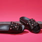 Black slides with upper made of goat leather with round metal studs over pink background.