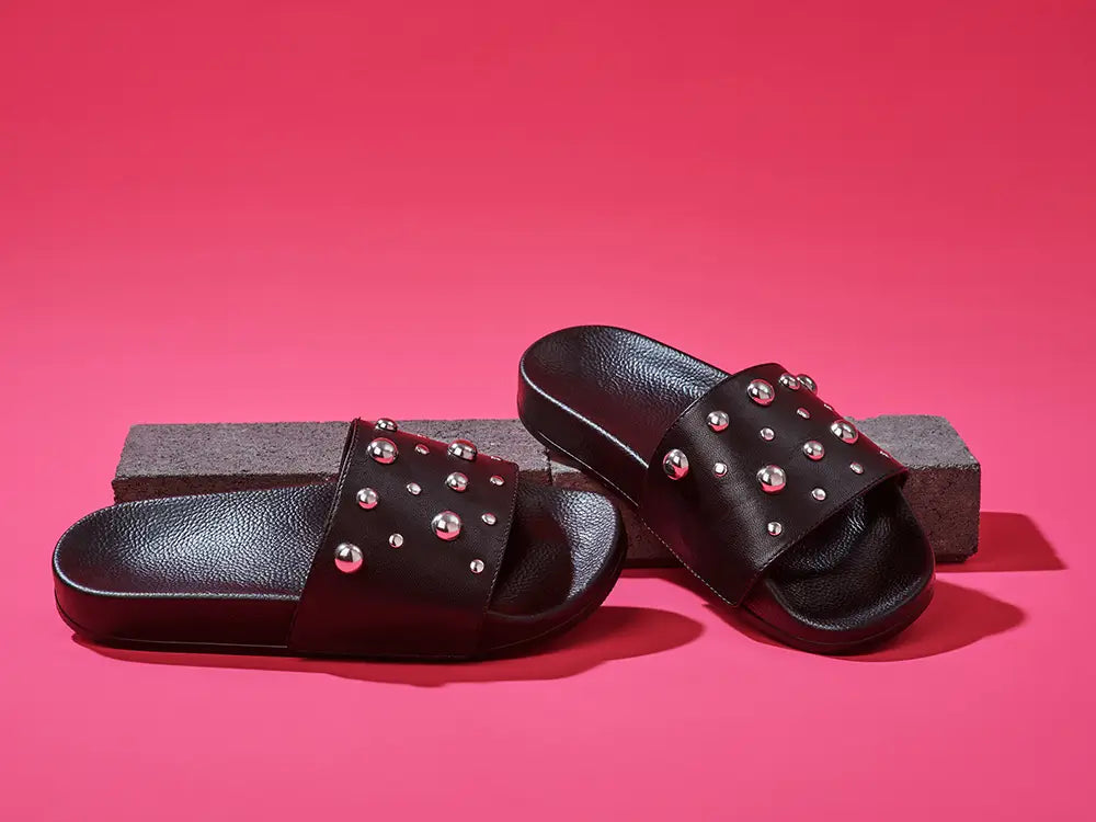 Black slides with upper made of goat leather with round metal studs over pink background.